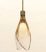 A modern pendant light with a clear glass teardrop-shaped shade featuring organic brown swirls, illuminated with a warm yellow glow, suspended from a brass-colored rod against a light beige background.
