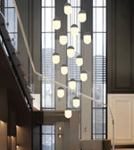 A modern multi-light pendant chandelier with multiple frosted glass dome bulbs suspended at varying heights by curved metal arms, shown hanging in a contemporary indoor staircase lobby.