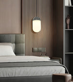 A modern bedroom scene featuring a wall-mounted cylindrical wall lamp with a glowing frosted white shade and a dark gray top, set against a brown vertical slat accent wall above a bed.