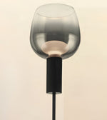 A modern floor lamp featuring a large, translucent smoky gray glass shade and a ribbed black metal stem, shown against a plain light beige background.