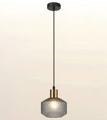 A modern single pendant light with a textured, faceted gray glass shade, a brass-finished metal collar, and a black suspension rod and ceiling canopy, shown against a soft beige gradient background.