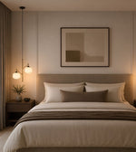 A modern bedroom featuring a neatly made bed with white and taupe bedding, a matching headboard, a framed abstract print on the wall, and warm pendant lights on a nightstand.