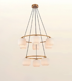 A modern multi-tier chandelier with five frosted glass shades and a brushed brass/gold metal frame, suspended by black cords against a plain off-white background.