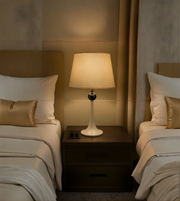 Two beds with white bedding and gold pillows, a nightstand with a lamp between them in a dimly lit room.