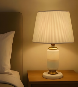 Table lamp with a white shade and gold base on a wooden nightstand next to a bed.