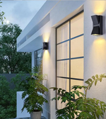 Modern outdoor wall lights on a house exterior with plants and trees in the background.