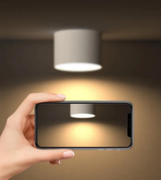 Hand holding a smartphone with a light fixture on the screen, against a blurred background.
