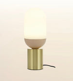 A modern table lamp with a glowing warm white capsule-shaped shade, a matte beige lower section, and a brushed gold cylindrical base, shown against a soft cream background.