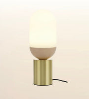 A modern table lamp with a glowing warm white capsule-shaped shade, a matte beige lower section, and a brushed gold cylindrical base, shown against a soft cream background.