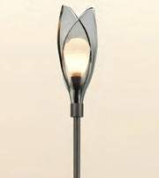 A modern tulip-shaped floor lamp with a glowing warm bulb inside a smoky translucent glass shade, mounted on a dark metal pole against a plain light beige background.