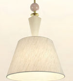 A modern pendant light fixture with a large tapered drum shade made of textured cream fabric, a white geometric stem, a pale pink spherical accent, and brass hardware against a plain off-white background.