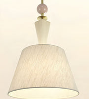 A modern pendant light fixture with a large tapered drum shade made of textured cream fabric, a white geometric stem, a pale pink spherical accent, and brass hardware against a plain off-white background.