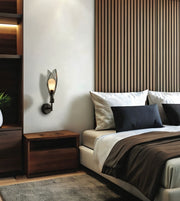 A modern wall-mounted sconce with a black metal frame and a warm glowing glass shade, mounted on a white wall in a contemporary bedroom next to a wooden nightstand and bed.