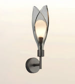 A modern wall sconce light fixture with a dark metal wall mount and a translucent smoky glass shade shaped like stylized leaves, shown illuminated against a soft beige background.
