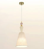 A modern pendant light with a tapered off-white fabric shade, a white glass geometric accent, a pale pink bead, and a gold-toned ceiling canopy and cord, shown against a plain off-white background.