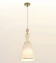 A modern pendant light with a tapered off-white fabric shade, a white glass geometric accent, a pale pink bead, and a gold-toned ceiling canopy and cord, shown against a plain off-white background.