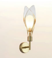A modern wall-mounted sconce with a warm brass-finished metal base and a clear glass shade shaped like stylized leaves, glowing with a warm light against a soft beige background.