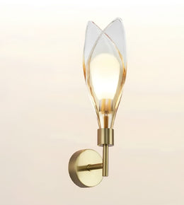 A modern wall-mounted sconce with a warm brass-finished metal base and a clear glass shade shaped like stylized leaves, glowing with a warm light against a soft beige background.