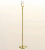 A modern floor lamp with a slim brass-colored metal pole, a round base, and a clear tulip-shaped glass shade that glows with a warm light, set against a soft beige gradient background.