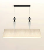 A modern rectangular pendant light fixture with a wide, tapered beige fabric shade, suspended from a black horizontal bar with two clear glass cone drops and dark gray spherical accents.