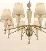 A modern six-light chandelier with a brushed brass/gold metal frame, cream fabric drum shades, and white geometric cone and bead accents, shown against a plain off-white background.