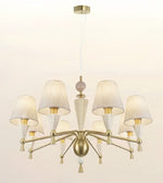 A modern six-light chandelier with a brushed brass/gold metal frame, white fabric drum shades, and clear glass cone accents, suspended from a matching ceiling canopy against a plain off-white background.
