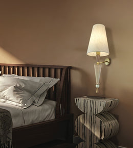A modern wall-mounted sconce with a white tapered shade, clear glass cone, and gold-toned metal arm, lit and mounted on a warm brown bedroom wall beside a dark wood bed and patterned side table.