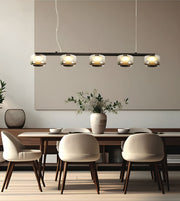 A modern 5-light linear pendant chandelier with clear glass shades and warm metallic accents, hanging over a dining table with cream upholstered chairs in a minimalist dining room.