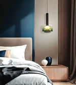 A modern pendant light with a black cylindrical shade and a green-tinted frosted glass globe bulb, glowing with a warm yellow light. It hangs in a contemporary bedroom above a wooden nightstand.