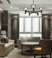 A modern living room interior featuring a multi-light chandelier with cylindrical glass shades, a light gray sofa, a cream sofa, a round wooden coffee table, and a brown armchair with a yellow cushion, set against dark wood paneling and sheer curtains.