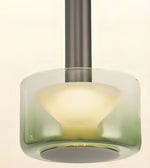 A modern pendant light with a cylindrical glass shade featuring a green-to-clear gradient and a frosted white inner diffuser, suspended by a dark gray metal rod against a warm off-white background.