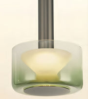 A modern pendant light with a cylindrical glass shade featuring a green-to-clear gradient and a frosted white inner diffuser, suspended by a dark gray metal rod against a warm off-white background.