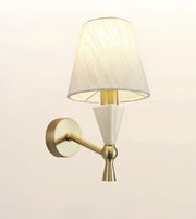 A modern wall-mounted sconce with a tapered off-white fabric shade, a white geometric cone detail, and a brushed gold metal arm and wall plate, shown against a plain light background.