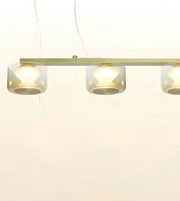 A modern linear pendant light fixture with three clear cylindrical glass shades and a brushed brass or gold-toned metal bar, shown against a plain white background.