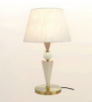 A modern table lamp with a tapered off-white fabric shade, a white spherical accent, a geometric off-white base, and a round gold metal base, shown against a plain light background.