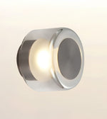 A modern cylindrical wall sconce with a polished chrome finish, emitting a warm light from its center against a soft beige gradient background.