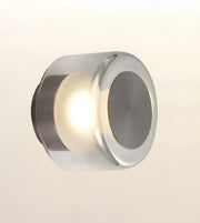 A modern cylindrical wall sconce with a polished chrome finish, emitting a warm light from its center against a soft beige gradient background.