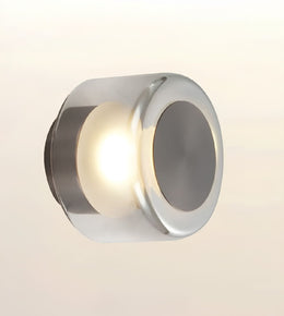 A modern cylindrical wall sconce with a polished chrome finish, emitting a warm light from its center against a soft beige gradient background.