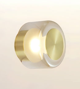 A modern cylindrical wall sconce with a brushed gold metal body, featuring a glowing warm white light in the center and a polished gold circular face on the right, set against a soft beige gradient background.