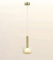 A modern single pendant light with a brushed gold metal cylinder stem and a frosted white glass shade, suspended by a thin cord against a warm off-white background.