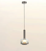 A modern pendant light fixture with a brushed metal cylindrical stem, a clear glass shade, and a glowing bulb inside, suspended by a thin cord from a matching ceiling canopy against a soft beige background.
