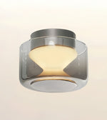 A modern semi-flush mount ceiling light fixture with a cylindrical chrome metal frame and a frosted glass shade that glows with a warm yellow light, shown against a plain off-white background.