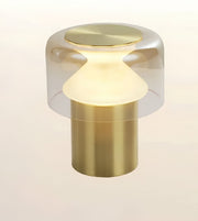 A modern table lamp with a brushed brass cylindrical base, a frosted white conical shade, and a clear glass dome top with a brushed brass lid, shown against a light beige background.