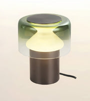 A modern table lamp with a translucent smoky green glass shade, a glowing warm white frosted center, and a matte dark bronze cylindrical base on a light beige background.