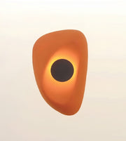 An abstract sculptural wall light or decorative object with a rounded organic shape in orange and yellow, featuring a dark circular center, shown against a plain light background.
