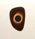 An abstract organic-shaped wall sconce or decorative light fixture with a dark brown outer body, a glowing warm orange ring, and a black circular center, shown against a plain light beige background.
