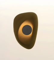 An abstract organic-shaped wall sconce or decorative light fixture with a textured metallic gold finish and a dark circular center, shown against a plain off-white background.