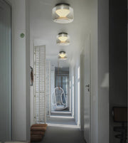 Three modern flush-mount drum ceiling lights with brushed silver metal tops and white shades, illuminated in a hallway with white walls and a hanging chair in the distance.