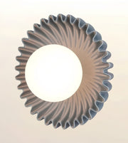 A modern circular pendant light with a wide, fluted scalloped rim and a central glowing white globe shade, shown against a plain off-white background.