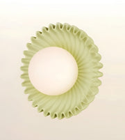A modern wall-mounted light fixture with a textured, fluted circular shade in a pale sage green and a glowing frosted white globe bulb, shown against a soft off-white background.
